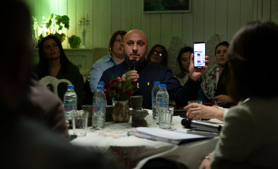 A person holding a phone in front of a group of people
Description automatically generated